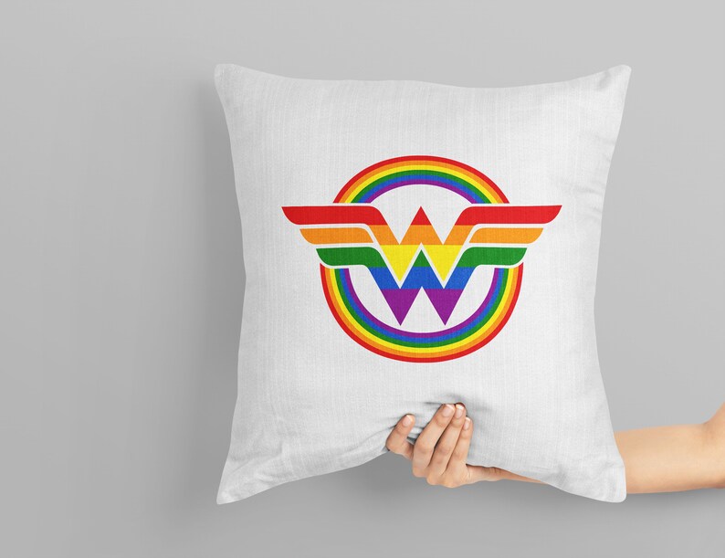 May include: A white pillow featuring a rainbow Wonder Woman logo. The logo is a circle with a rainbow design and a purple W at its centre. The wings are red, orange, yellow, green, blue, and purple.