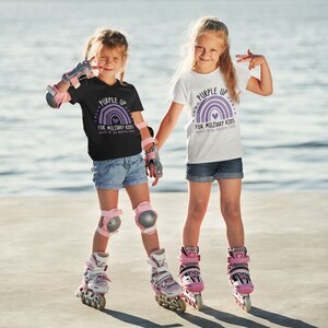 May include: Two children wearing 'Purple Up for Military Kids' t-shirts with a rainbow graphic and heart. The children are wearing rollerblades and protective gear.