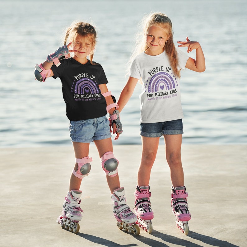 May include: Two children wearing 'Purple Up for Military Kids' t-shirts with a rainbow graphic and heart. The children are wearing rollerblades and protective gear.