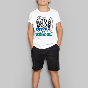 May include: A white t-shirt with a blue and black graphic that says "100 Days of School" with paw prints and pencils. The shirt is being worn by a child.