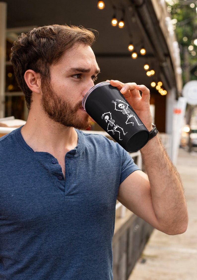 May include: A black travel mug with a white design of two skeletons dancing. The mug is made of stainless steel and has a clear lid.