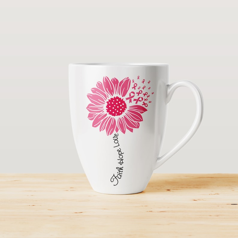 May include: White ceramic coffee mug with a pink sunflower design. The sunflower has pink ribbon designs around it. The text "Faith Hope Love" is written vertically on the mug.