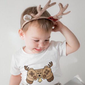 May include: A toddler wearing a white t-shirt with a brown video game controller graphic and a reindeer headband with glitter and a red bow. The toddler is sitting down and looking down.
