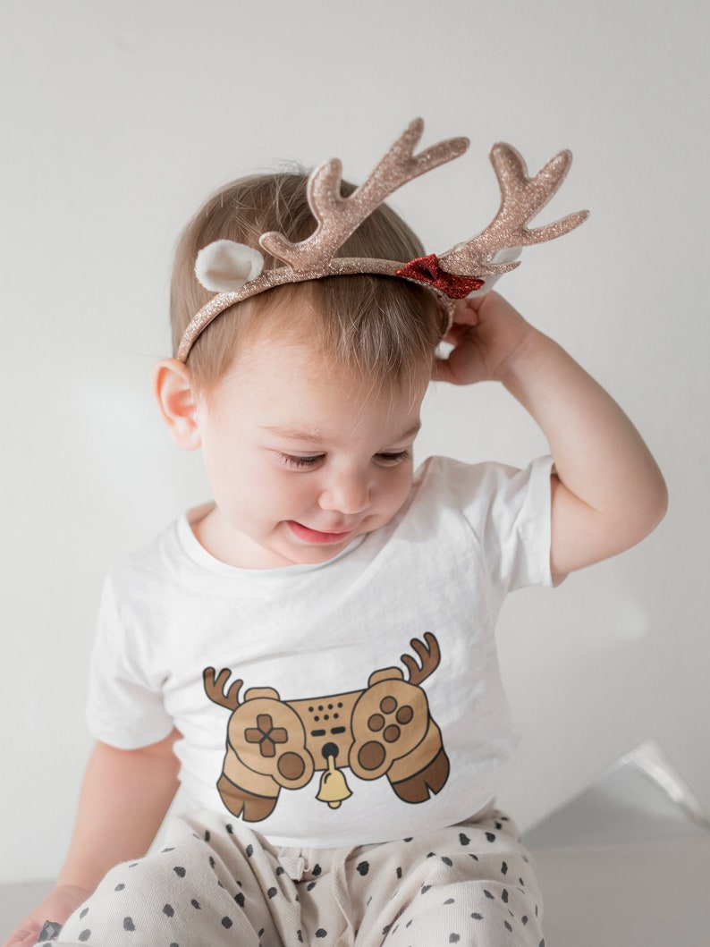 May include: A toddler wearing a white t-shirt with a brown video game controller graphic and a reindeer headband with glitter and a red bow. The toddler is sitting down and looking down.
