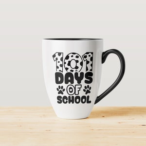 K&ouml;nnte beinhalten: Wei&szlig;e Keramiktasse mit schwarzem Henkel. Die Tasse hat ein schwarzes Design mit der Aufschrift "101 Tage Schule" mit Pfotenabdr&uuml;cken und einem Herzen.