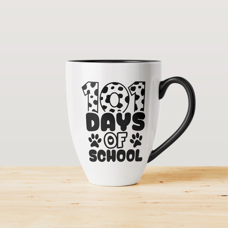 K&ouml;nnte beinhalten: Wei&szlig;e Keramiktasse mit schwarzem Henkel. Die Tasse hat ein schwarzes Design mit der Aufschrift "101 Tage Schule" mit Pfotenabdr&uuml;cken und einem Herzen.