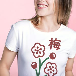 May include: White t-shirt featuring a floral design with the Japanese character for plum. The design includes maroon flowers, berries, and green stems. The shirt is a classic crew neck style.