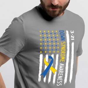 Może przedstawiać: Szary T-shirt z wzorem biało-niebieskiej flagi amerykańskiej. Flaga ma ż&oacute;łto-niebieską wstążkę w środku i napis "3-21 DOWN SYNDROME AWARENESS" po prawej stronie.
