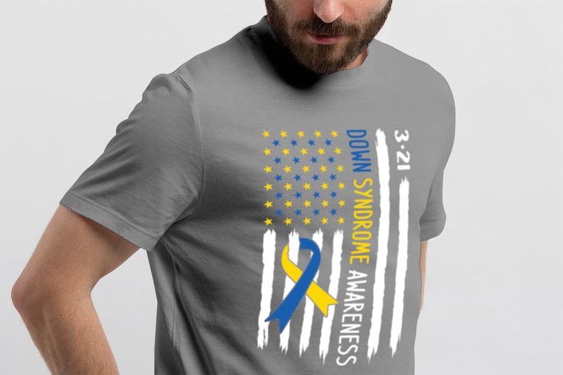 Może przedstawiać: Szary T-shirt z wzorem biało-niebieskiej flagi amerykańskiej. Flaga ma ż&oacute;łto-niebieską wstążkę w środku i napis "3-21 DOWN SYNDROME AWARENESS" po prawej stronie.