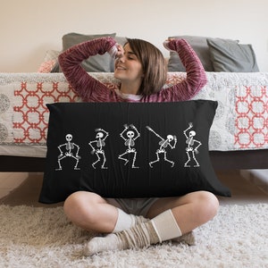 May include: A black throw pillow with a white design of five skeletons dancing. The skeletons are in various poses, with their arms and legs outstretched. The pillow is sitting on a bed with a red and white patterned comforter.