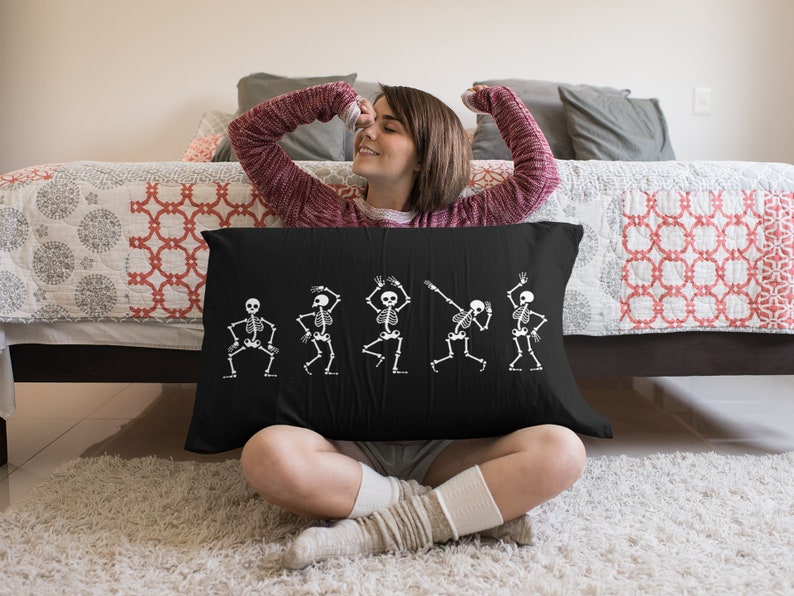 May include: A black throw pillow with a white design of five skeletons dancing. The skeletons are in various poses, with their arms and legs outstretched. The pillow is sitting on a bed with a red and white patterned comforter.