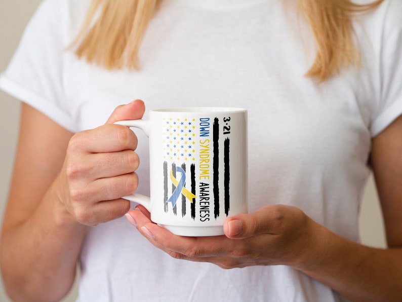 Może przedstawiać: Biały kubek ceramiczny z niebieską i ż&oacute;łtą wstążką i tekstem "Down Syndrome Awareness 3-21" na czarno-białym wzorze amerykańskiej flagi.