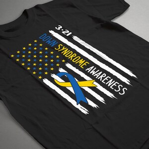 Może przedstawiać: Czarny T-shirt z wzorem biało-niebieskiej flagi amerykańskiej. Flaga ma ż&oacute;łto-niebieską wstążkę w środku i napis "3.21 Down Syndrome Awareness".