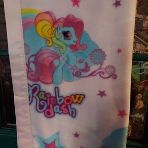 May include: Pink fleece blanket with a blue and rainbow cartoon pony with a rainbow mane and tail. The blanket has a white background with stars and the words "Rainbow Dash" in a colorful font.