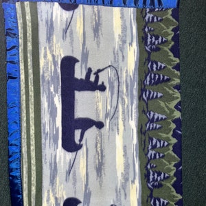 May include: A blue and green fleece blanket with a design of two people fishing in a canoe on a lake. The blanket has a blue satin trim around the edges.