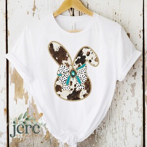 May include: White t-shirt with a brown and white cowhide bunny design with a turquoise bow and a jewel accent.