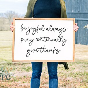 May include: A white wooden sign with black calligraphy that reads "be joyful always pray continually give thanks".