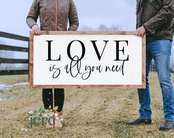 May include: A couple holds a wooden sign with the text "LOVE is all you need" in black lettering. The sign is white with a brown wooden frame. The couple is standing in a snowy field with a wooden fence in the background.