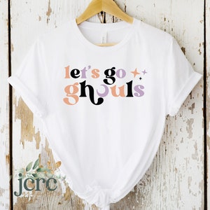 Könnte beinhalten: Weißes T-Shirt mit dem Schriftzug "let's go ghouls" in einem Retro-Schriftstil. Der Text ist in Pfirsich-, Schwarz-, Lavendel- und Orangetönen gehalten. Das Shirt hängt an einem Holzkleiderbügel vor einem weißen Holzgrund.