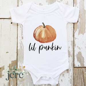 May include: White baby onesie with a watercolor pumpkin and the text "lil pumpkin" in black.