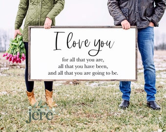 May include: A white wooden sign with the text "I love you for all that you are, all that you have been, and all that you are going to be." The sign is being held by two people in a field.