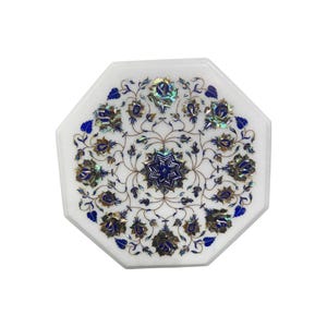 Puede incluir: Tablero de mesa octogonal de mármol blanco con incrustaciones florales. El diseño presenta detalles azules e iridiscentes, creando un patrón detallado. Este tablero decorativo es adecuado para varios estilos de decoración del hogar.