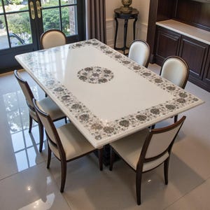 Puede incluir: Una mesa de comedor rectangular de mármol blanco con un borde floral y un medallón central. Seis sillas con asientos tapizados de color crema y marcos de madera oscura rodean la mesa. La habitación tiene un suelo pulido.