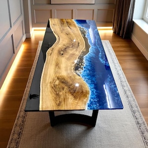 May include: A handcrafted wooden dining table with a unique design. The table features a live edge wood center, black resin on one side, and a blue resin ocean design on the other. The table is supported by a black base.