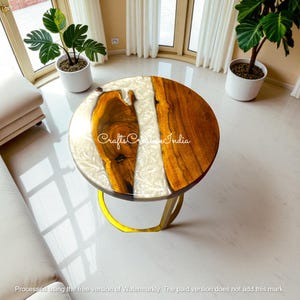 Puede incluir: Una mesa de centro redonda con una tapa de madera y una incrustación de resina blanca. La mesa tiene una base de metal amarillo. El texto "Crafts Creative India" es visible en la superficie de la mesa.