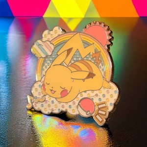 May include: Gold-tone enamel pin featuring a sleeping Pikachu on a cloud with a rainbow and a sun. The pin has a colorful, dotted background.