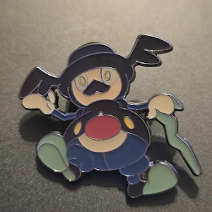 May include: Enamel pin featuring a cartoon character with a black hat, mustache, and blue outfit. The pin has a red belly, yellow accents, and green shoes. The character is holding a green staff.