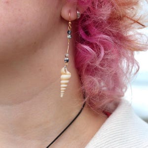 Könnte beinhalten: Ein goldfarbener Drahtohrring mit einem Muschelanhänger. Der Ohrring hat kleine, dunkle Perlen und eine spiralförmige Muschel. Das Haar der Person ist rosa und orange.
