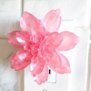 May include: A pink flower-shaped plug-in air freshener. The flower is made of a translucent material with a shimmery finish.