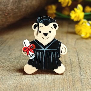 May include: A wooden bear figurine wearing a black graduation gown and cap, holding a scroll with a red ribbon. The bear has a brown nose and black eyes.