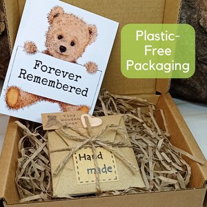May include: A small, brown wooden bear with a heart-shaped tag that says "Tiny Wooden Bear" is packaged in a brown paper envelope with a twine bow and a "Hand Made" tag. The envelope is sitting on a bed of shredded paper inside a cardboard box. The box has a green label that says "Plastic-Free Packaging". A card with a cartoon bear holding a sign that says "Forever Remembered" is also included.
