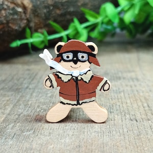 Peut inclure: Un ours en bois jouet portant une veste d'aviateur marron et des lunettes. L'ours a une écharpe blanche et est peint avec des détails marrons, blancs et noirs.