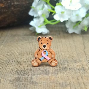 May include: Wooden bear figurine holding a pink and blue ribbon. The bear is brown and has a painted face. The ribbon is a symbol of awareness for a cause.