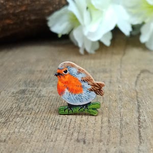 May include: A wooden robin bird figurine with a bright orange breast, grey and brown wings, and a green perch. The bird is perched on a green branch, set against a blurred wooden background.