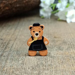 May include: A wooden bear-shaped pin with a black apron and hat, holding a yellow spoon. The bear is light brown with dark brown details. The pin is on a wooden surface.