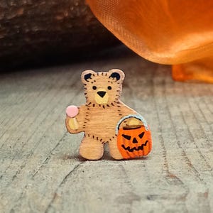 May include: A small, flat, light brown wooden bear cutout featuring a painted black nose, ears, and stitched details. The bear holds a pink lollipop in one hand and carries an orange jack-o'-lantern pumpkin bucket with a silver handle.