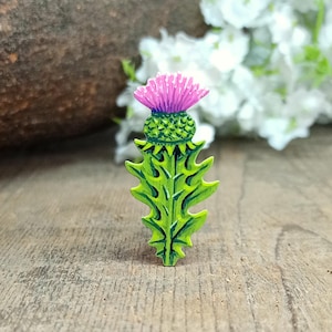 以下が含まれることがあります： 鮮やかな紫色の花頭、質感のある緑色の基部、そしてギザギザした尖った緑色の葉の茎が特徴のアザミのピン。このデザインは詳細な陰影と明確な線を表示し、植物の植物学的構造を平らで直立した向きで捉えています。