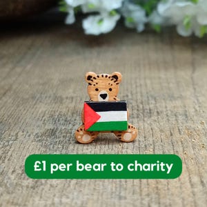 May include: Wooden bear figurine holding a Palestinian flag. The bear is painted brown and has a black nose and eyes. The flag is red, black, white, and green. The text "£1 per bear to charity" is printed on a green banner below the bear.