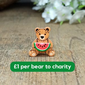 May include: Wooden bear figurine holding a slice of watermelon. The bear is painted brown with black spots and has a red nose. The watermelon is painted red with green rind. The text "£1 per bear to charity" is printed on a green banner below the bear.