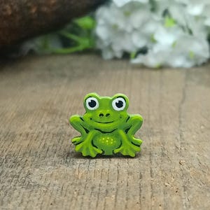 May include: A small, green frog-shaped pin with a smiling face and white eyes. The pin is painted with a light green body and darker green outlines. The pin is on a wooden surface.