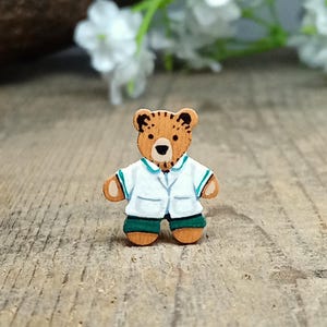 May include: A wooden bear brooch with a light brown body, dressed in a white coat with a teal collar and green shorts. The bear has a black nose and eyes. The brooch is set against a blurred background of white flowers and a wooden surface.