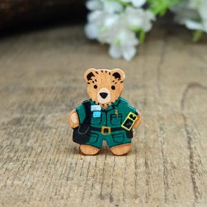 May include: A wooden bear figurine wearing a green uniform with a black bag and a yellow badge. The bear has brown fur and a black nose.