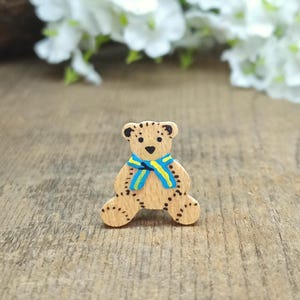 May include: A small wooden teddy bear figurine with a blue and yellow striped scarf. The bear is painted brown with black accents and has a simple, hand-painted design.