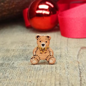 May include: A small, light brown wooden bear-shaped button featuring dark brown spots, a black heart-shaped nose, and a gold-colored bow tie detail at the neck. The bear has a smooth finish and is designed with a flat surface for attachment to garments.