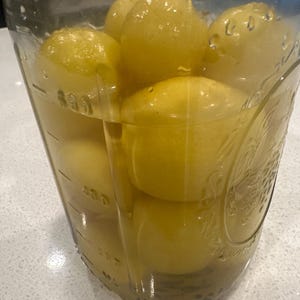May include: A clear glass jar filled with yellow lemons submerged in a liquid. The jar has measurement markings on the side. The lemons are tightly packed, and the liquid appears slightly cloudy.