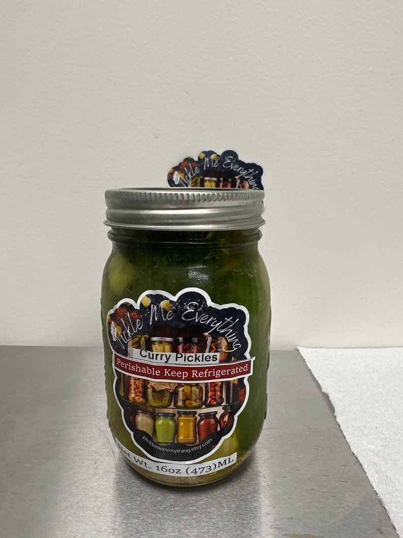 K&ouml;nnte beinhalten: Ein Glas mit gr&uuml;nen Curry-Pickles. Das Glas hat einen silbernen Deckel und ein Etikett mit der Aufschrift "Curry Pickles" und "Perishable Keep Refrigerated". Das Etikett zeigt auch eine Collage von Gl&auml;sern und den Text "Pickle Me Everything."